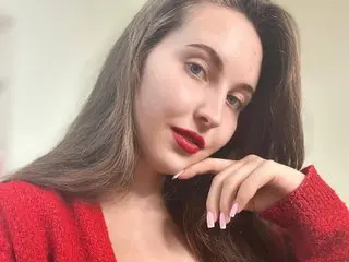 Kornika poses with long brown hair and red lipstick, wearing a red knit sweater against a plain light background.