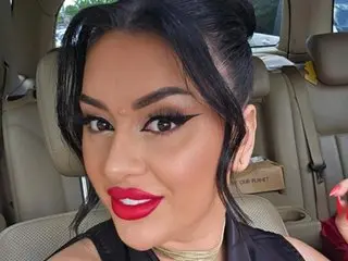 KristallMadness poses close-up with her black hair pulled back, wearing red lipstick and a black top while sitting in a car seat.