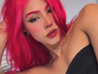 KristensClark poses with her bright pink hair and red lipstick, wearing a black top against a plain light background.
