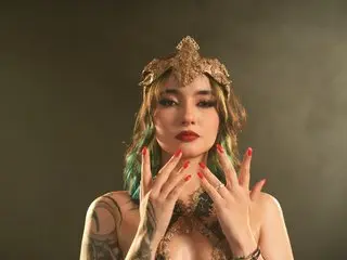 KyRoseuwu poses with her hands near her face, wearing a gold headpiece and a gold bra, with long dark hair and red lipstick.