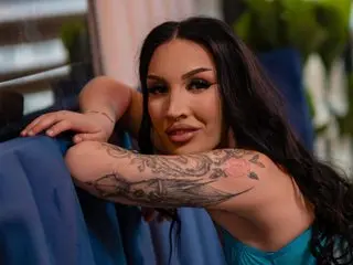 KyraReign poses with her long dark hair, showing her bare shoulders and chest while leaning forward against a blue surface.