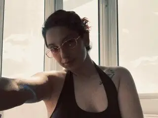 LEILAAx poses in a black tank top with her dark hair pulled back, looking toward the camera against a bright window background.