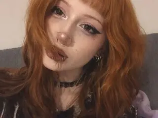 LIYNA poses close-up, showing her face and long, wavy red hair with blunt bangs, wearing a black choker and a dark patterned top.