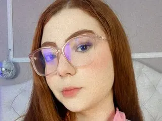 Young woman with red hair and clear glasses wearing pink clothing in a soft, bright indoor setting.