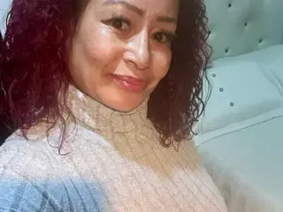 LIZZY49 poses close-up wearing a light tan turtleneck, showing her face and curly dark hair against a white tufted headboard.