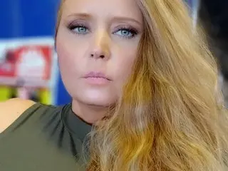 Lacii_lo poses with her long blonde hair framing her face, wearing a dark olive green top against a soft blue background.