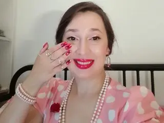 LadyDaphnne smiles while wearing a pink polka-dot top and a white pearl necklace, holding her hand near her face with red manicured nails.