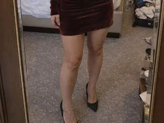 LadyLaraX stands in a room wearing a short red velvet dress and black heels, showing off her bare legs.
