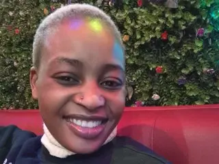 Smiling Black woman in a black sweater sitting on a red sofa against a lush green floral wall background.