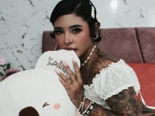 Tattooed woman in a white lace top holding a white plushie while sitting on a bed in a cozy room.