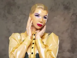 LatexRapture poses with her hands against her face, wearing a shiny tan latex top and heavy eye makeup with her hair slicked back.