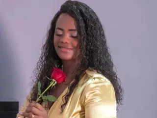 Smiling woman in a gold satin dress holding a single red rose against a plain background.
