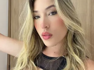 Blonde woman in a black mesh top posing with soft makeup in a bright indoor setting.