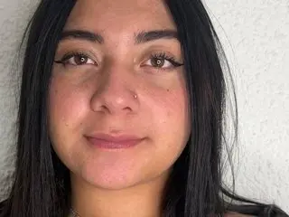 Close-up portrait of a smiling young woman with long dark hair and natural makeup against a white wall.
