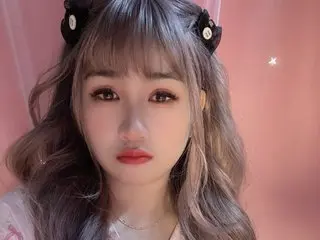LavenderBae looks at the camera with long wavy hair and bangs against a pink background.