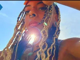 Leafgirl poses with her blonde-tipped braids, showing her face and chest against a bright blue sky background.