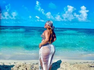 LeahxBarker poses on a beach wearing a white lace bikini, showing her bare back and curves against the blue ocean and sky.