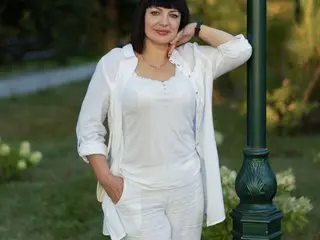 Smiling woman in elegant white linen outfit leaning against a green lamp post in a sunny park setting.