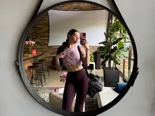 LennonTheAmazonian poses in a light purple bra and matching leggings, showing her hair down in a bright room with indoor plants.