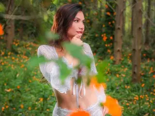 LeticiaHinkley poses with her hand under her chin, wearing a white lace bra or top against a soft, leafy outdoor background.
