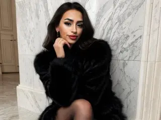 LexaSawyer poses with her hand under her chin, wearing a black furry coat and showing her long dark hair against a white marble wall.