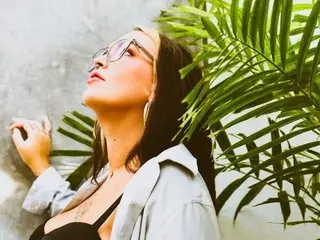 LexiLotusUS poses with her head tilted back, wearing glasses and a light blue button-down shirt against a light background with green palm leaves.