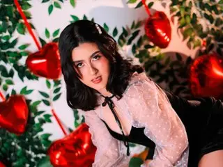 LiLLiAnderson poses in a black lace top, showing her dark hair and cleavage against a floral background.