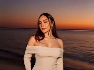 LiaBalden poses with long dark hair, wearing a white off-the-shoulder top against a sunset beach background.