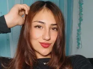 LiahLesley poses with her hand in her long brown hair, wearing a black top against a blue curtain background.