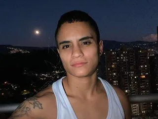 Liiam_thompson20 wears a white tank top and smiles outdoors at night with city lights in the background.