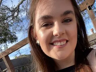 LilianRose33 smiles while looking at the camera, showing her face and long brown hair against a background of trees and a wooden fence.