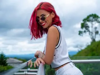 LilithLuxxor smiles while posing outdoors, wearing a white sleeveless top and sunglasses with her short red hair styled down.