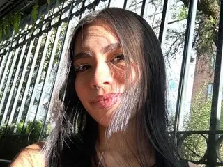 Young woman with long dark hair smiling in sunlight against a metal fence background.