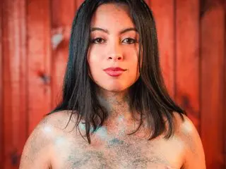 LilithSant is a brunette woman with long dark hair posing shirtless against a red wooden wall with blue glitter on her skin.