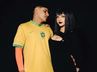 LilithStonne poses in a yellow t-shirt with black hair and dark makeup against a black background.
