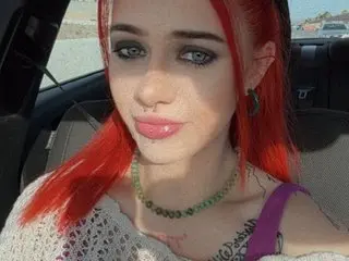 LillithSapphire poses in a car, showing her long red hair, light eyes, and a light lace top with a purple strap visible.