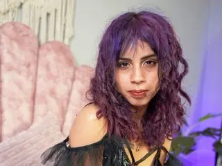 Liltrixxxy with purple hair wearing a black lace top, posing against a pink scalloped chair.