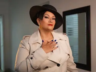 LilyanaJaramillo poses in a cream trench coat with her hands on her chest, showing her dark hair and red lipstick against a neutral background.