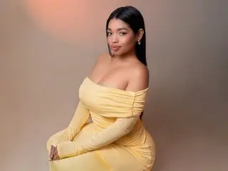 LilyyFox poses in a yellow off-the-shoulder dress, showing her bare shoulders and long dark hair against a plain background.