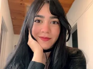 Lilyyy_Lily poses with her hands near her chin, showing her long black hair and a black choker against a plain indoor background.