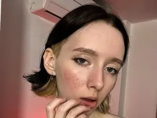 Close-up portrait of a young woman with dark hair and natural makeup in a soft pink indoor setting.