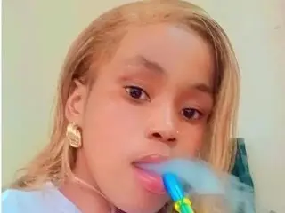 Lindabee29 poses with blonde hair and a blue straw in her mouth, looking directly at the camera against a plain wall.