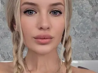 Lindy18 poses head-on with her blonde hair in two long braids, looking directly at the camera against a light grey patterned wall.