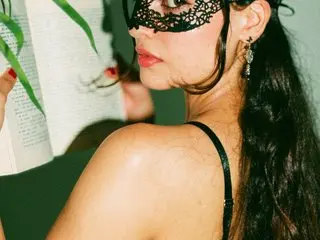 Mysterious woman in a black lace masquerade mask and black strap outfit reading a book in moody lighting.