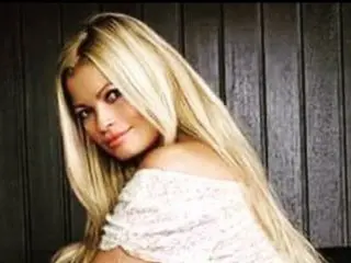LisaVonTease poses with her long blonde hair down, wearing a white lace off-the-shoulder top against a dark wooden background.