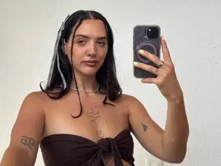 Young woman with braided hair in a brown strapless top taking a casual selfie against a white wall.