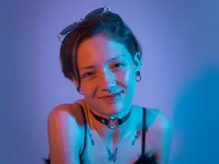 LissyLuxe smiles while wearing a black choker and a dark top, looking directly at the camera against a blue background.