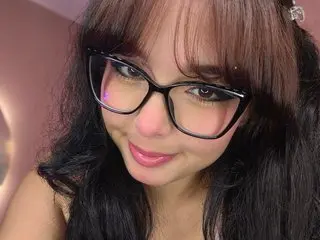 Smiling young woman with dark hair and black cat-eye glasses in a warm indoor setting.