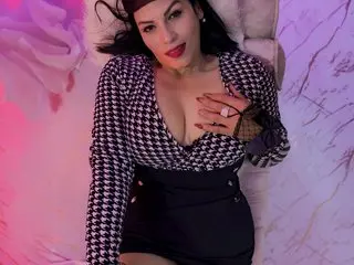 LizaRussell wears a black and white patterned top and black skirt while posing on a white surface.