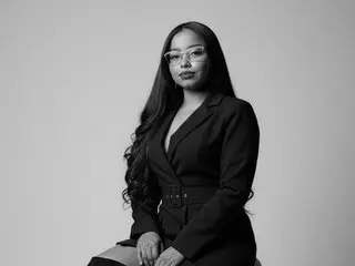 Lizstone26 poses in a black blazer and matching skirt, looking at the camera with long dark hair against a plain grey background.